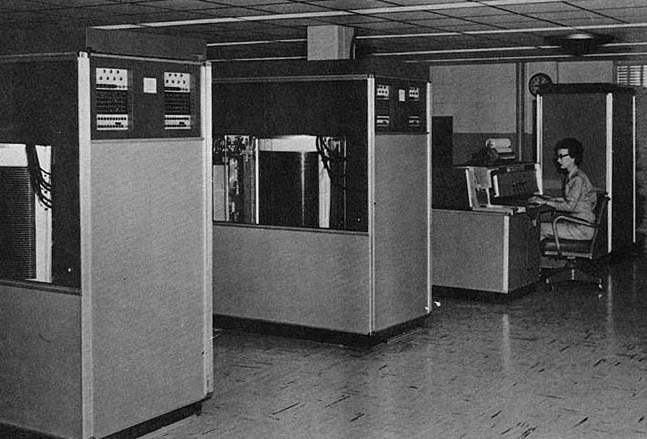 The IBM 305 RAMAC. In the fore- and midground of the photo are two big cabinets, about 1m wide by 3m deep by 2m tall. In the background is a woman sitting at the console, operating the computer.